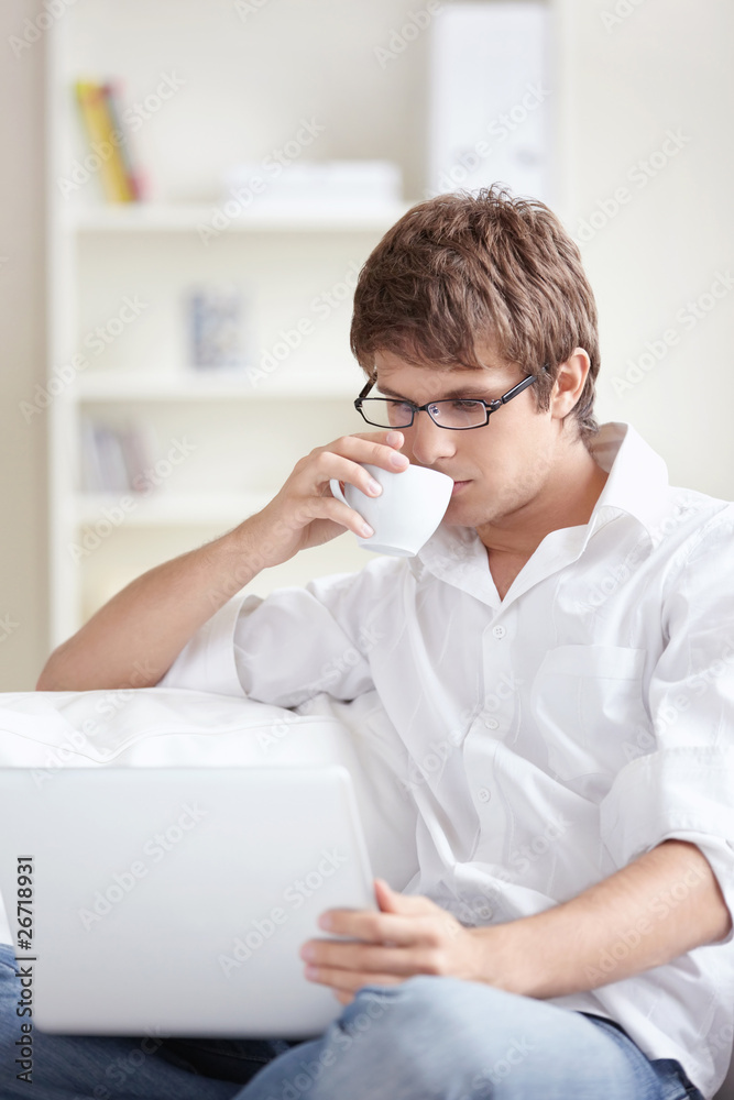 A young man with a cup of looking at the laptop at home