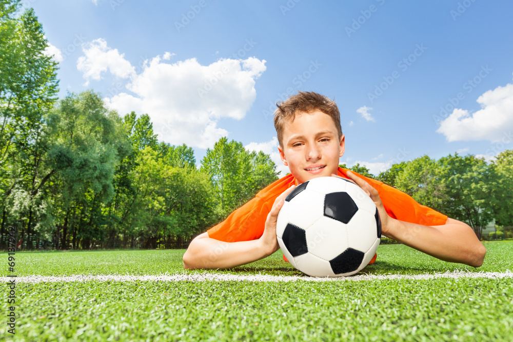 Happy boy holding football lays on green grass