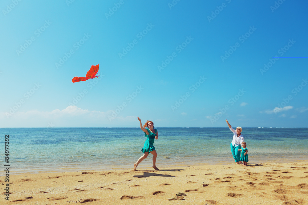 Carefree mother having a fun running with flying red kite on sea beach to baby girl and grandmother.