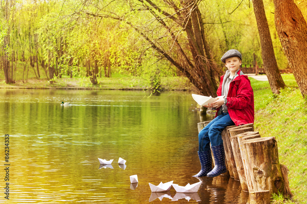 Boy sit near pond playing with white paper boats