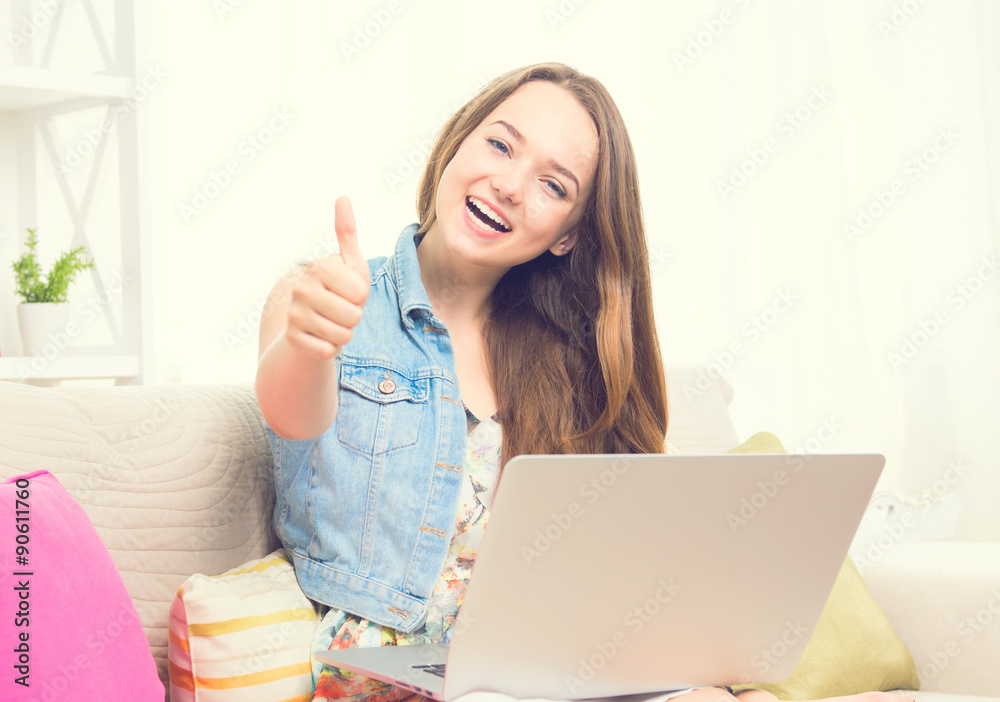 Pretty teenage girl sitting on sofa at home with her laptop, making thumb up