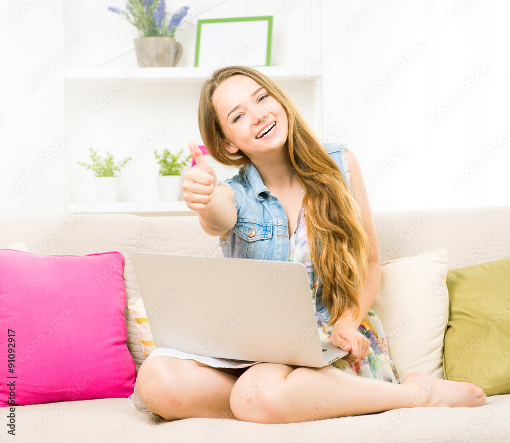 Beauty teenage girl sitting on sofa, using laptop and smiling. Showing thumbs up