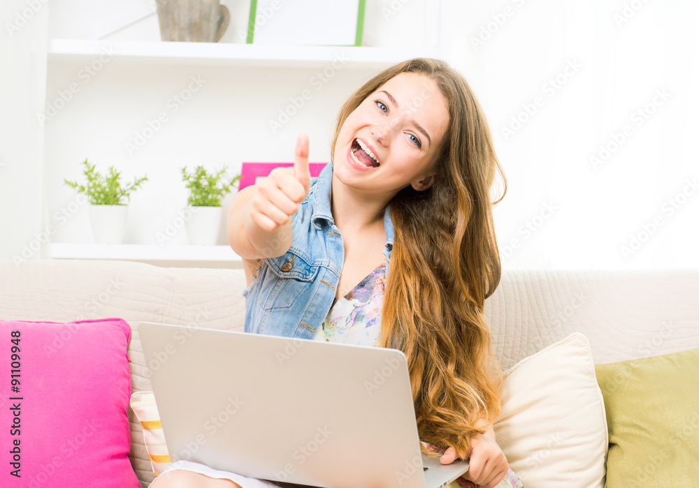 Beauty teenage girl sitting on sofa, using laptop and smiling. Showing thumbs up