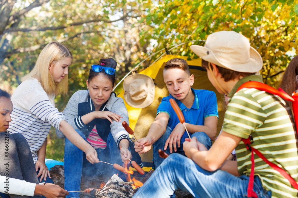 Teenagers on campsite grill sausages near bonfire