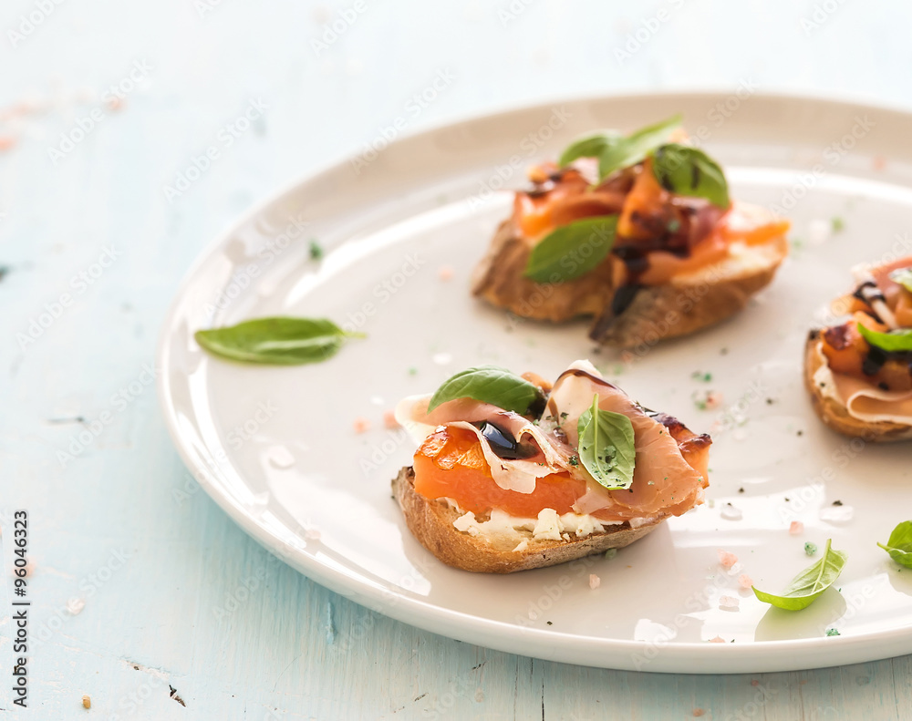 Bruschettas with Prosciutto, roasted melon, soft cheese and basil on white ceramic plate over light 