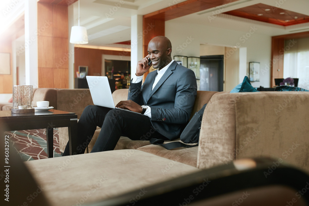 Happy businessman in hotel lobby using cell phone and laptop