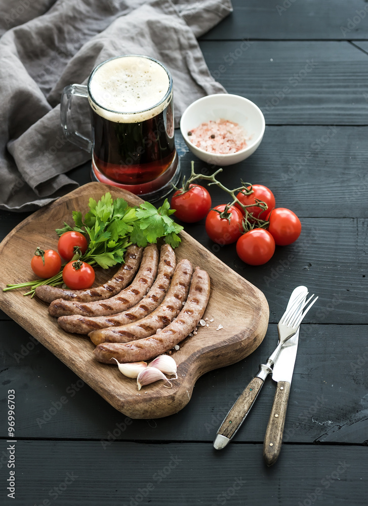 Grilled sausages with vegetables on rustic serving board and mug of light beer over black wooden bac