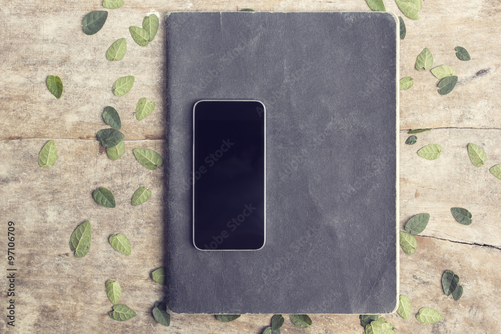 Blank black smartphone with exercise book and leaves on wooden t