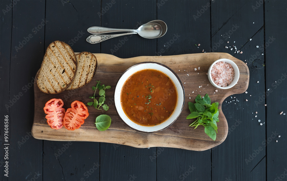 Roasted tomato soup with fresh basil, spices and bread in vintage metal bowl on wooden board over bl