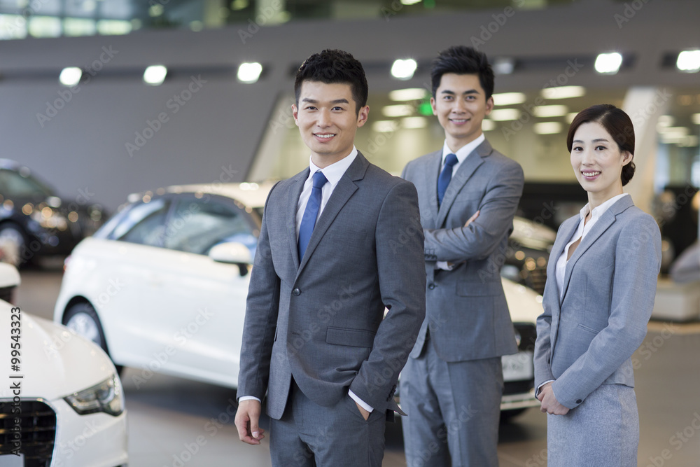 Confident salespeople standing with new cars in showroom