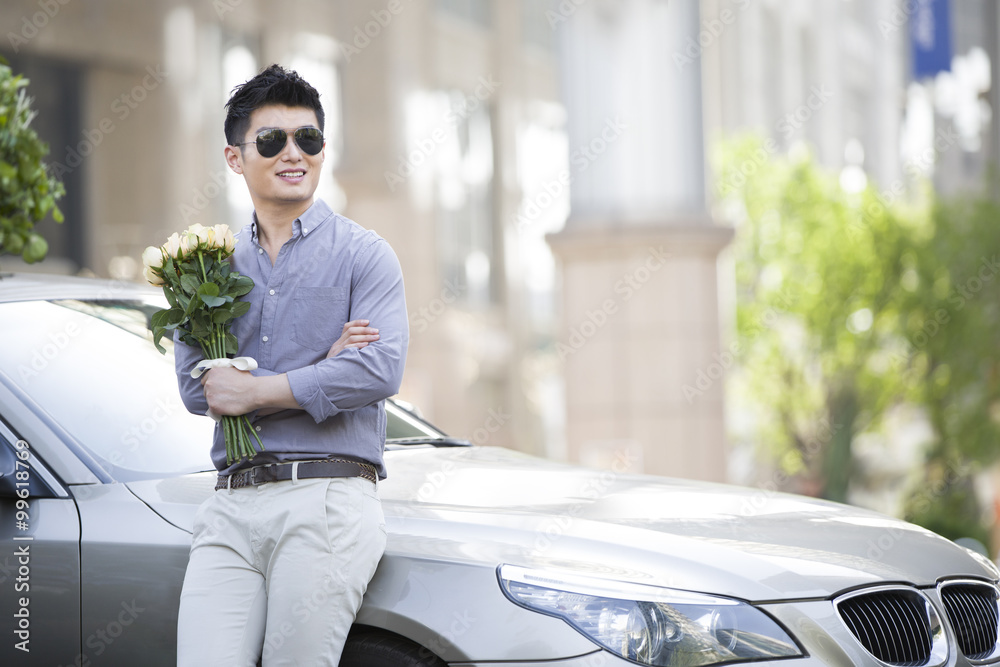 Young man leaning against his car with a bunch of flowers