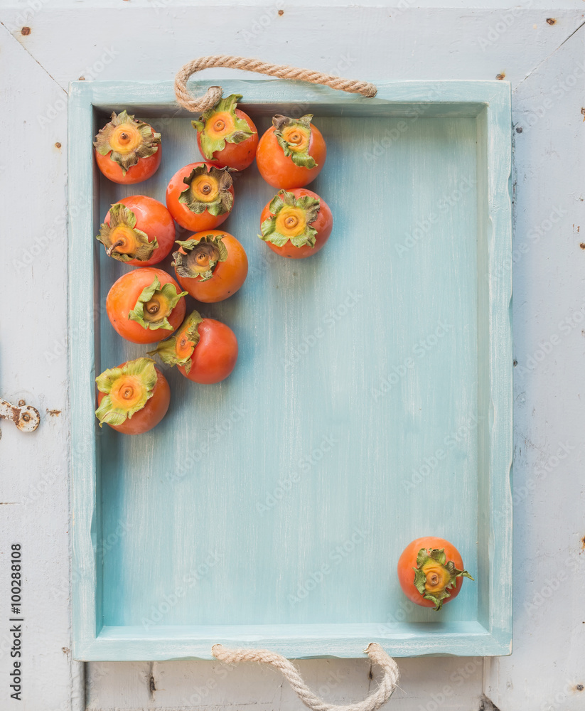 Ripe fresh persimmons on blue wooden tray over light backdrop, top view