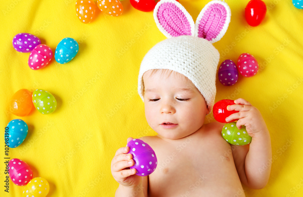 Baby boy in bunny hat lying on yellow blanket with easter eggs