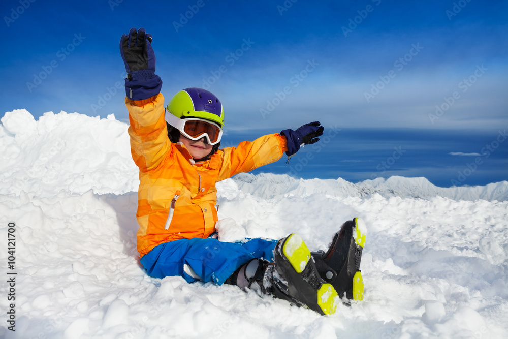 快乐的滑雪者男孩双手高举坐在雪地里