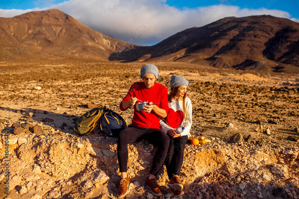 Young couple travelers eating healthy food from cooking pan sitting on the ground on desert mountain