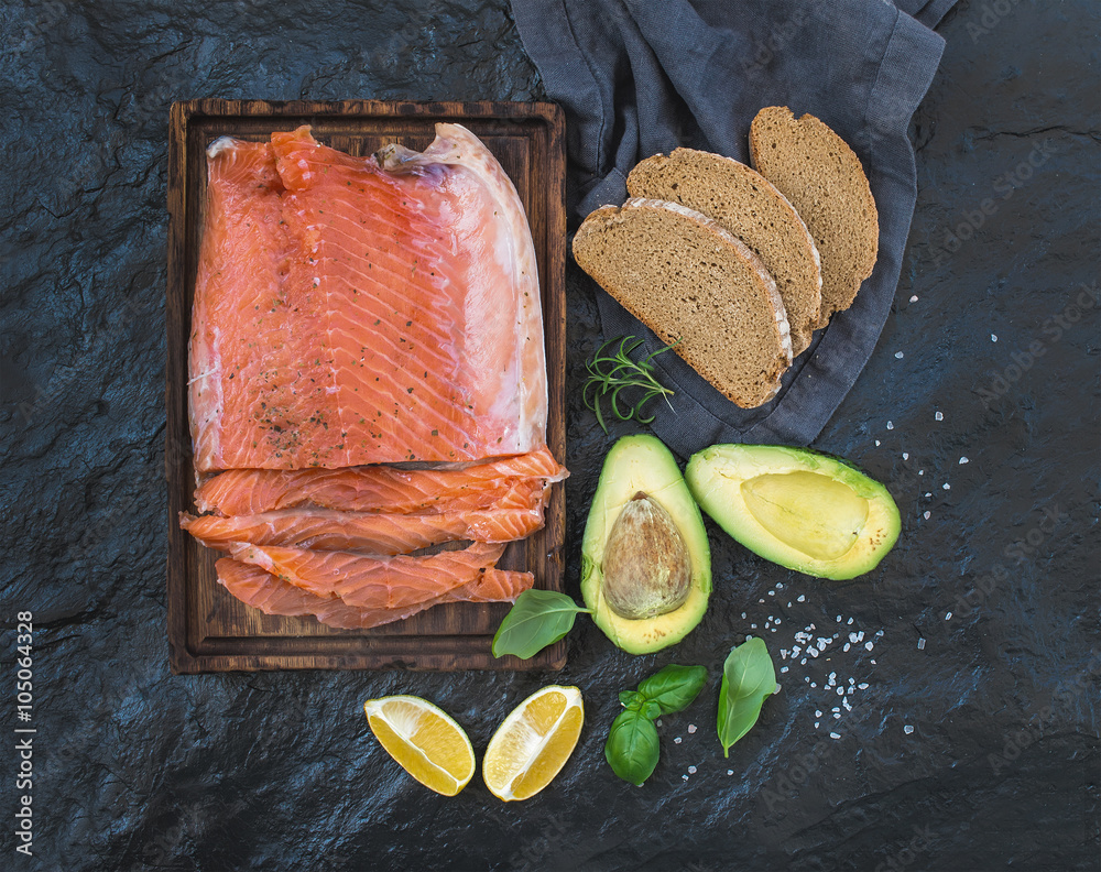 Smoked salmon filet with lemon, avocado, fresh herbs and bred on wooden serving board over dark ston