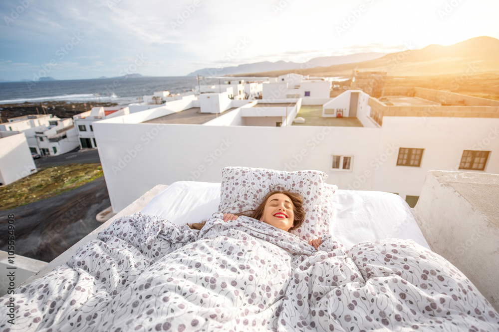 Young and cute woman sleeping on the bed on the roof top with white houses on the background. Concep