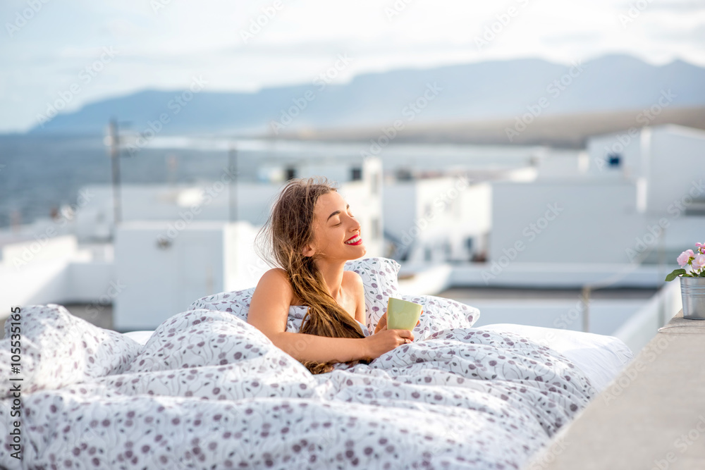 年轻可爱的女人拿着茶杯躺在屋顶的床上，白色的房子和山脉o