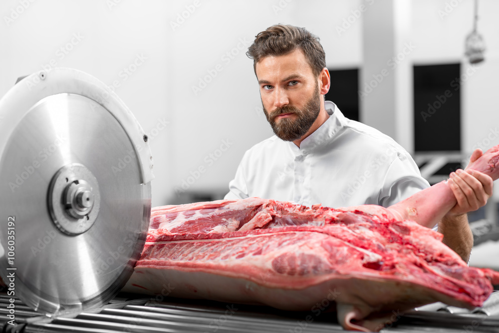 Handsome butcher cutting pork carcasse with big electric saw at the meat manufacturing