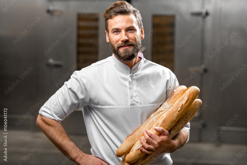 Handsome baker in uniform holding baguettes with oven on the background at the manufacturing