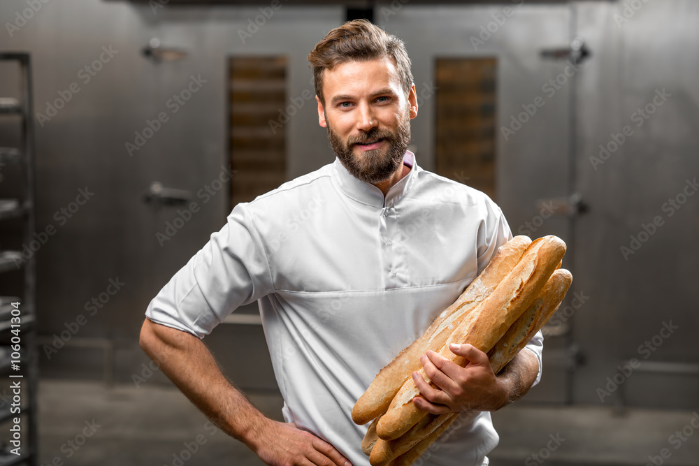 Handsome baker in uniform holding baguettes with oven on the background at the manufacturing