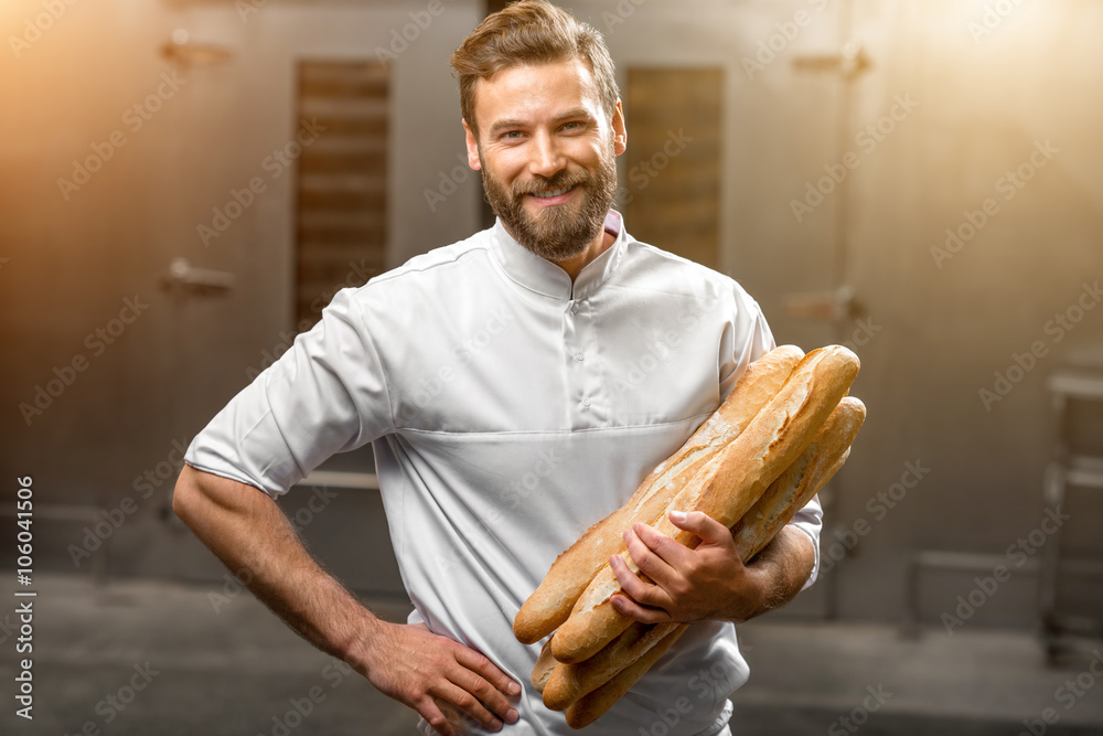 Handsome baker in uniform holding baguettes with oven on the background at the manufacturing