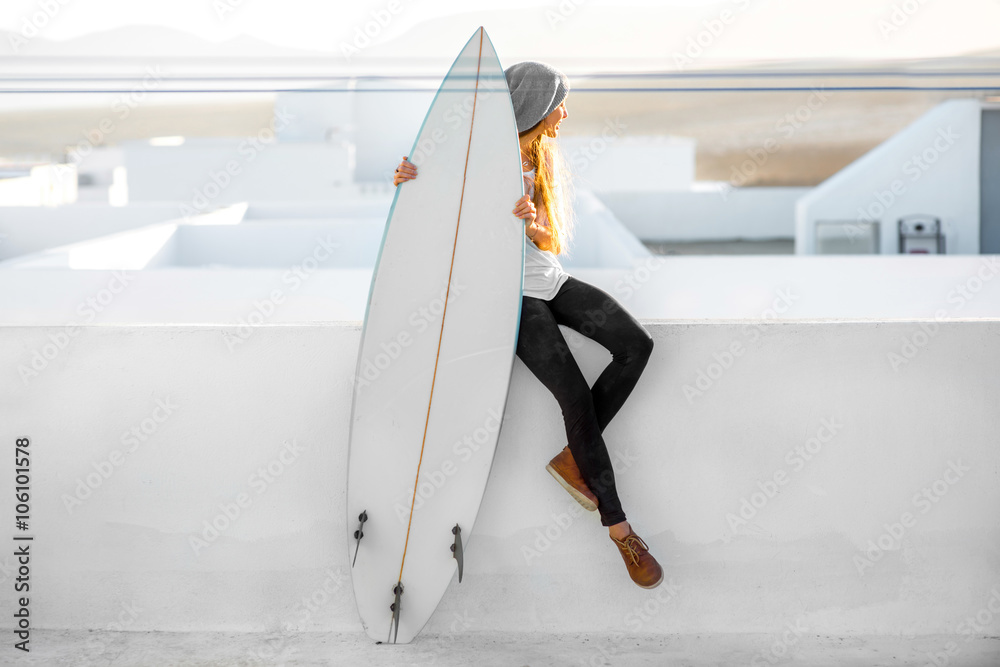 Young smiling woman in white t-shirt and hat sitting with surfboard on the roof top on the white cit