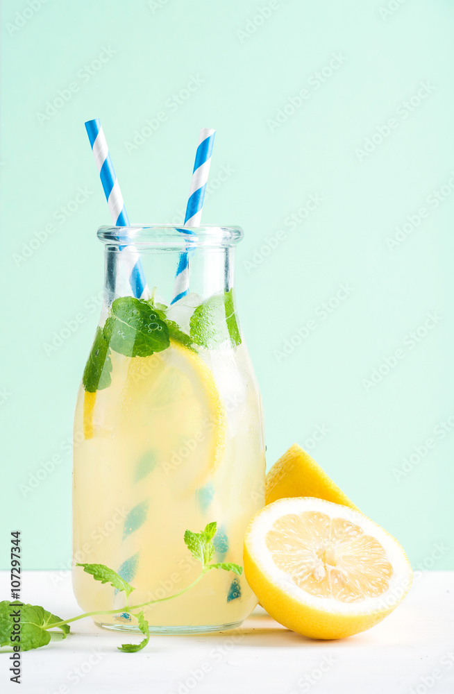 Bottle of homemade lemonade with mint, ice, lemons, paper straws and pastel blue background