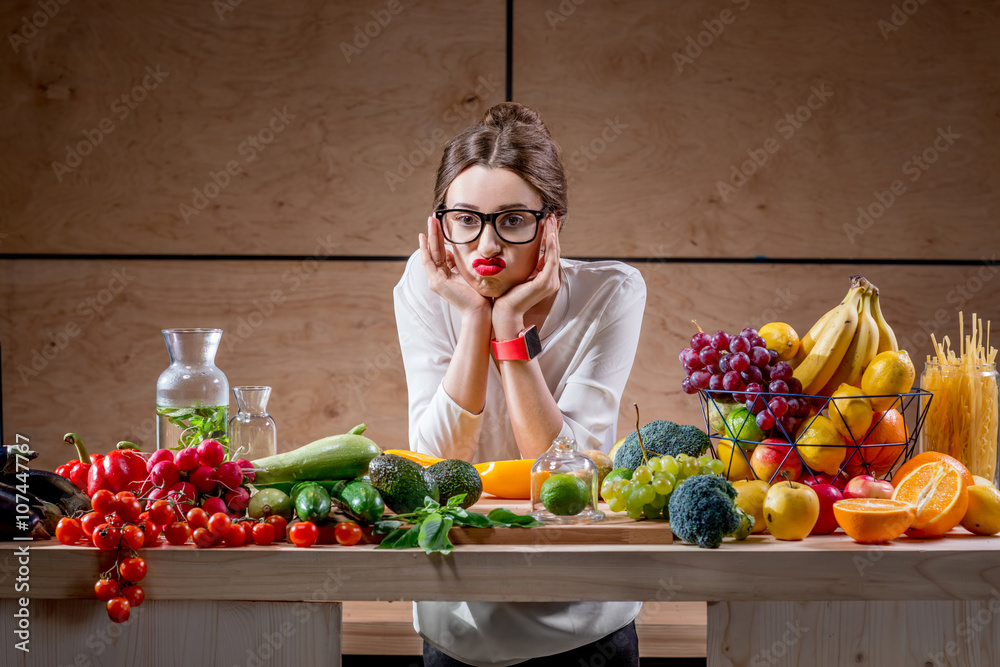 Young and sad woman thinking about tasty food and calories sitting at the table full of fruits and v