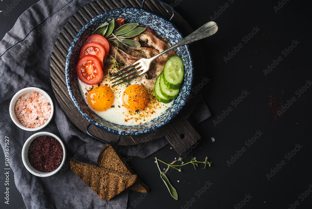 Breakfast set. Pan of fried eggs with bacon, fresh tomato, cucumber, sage and bread on dark serving 