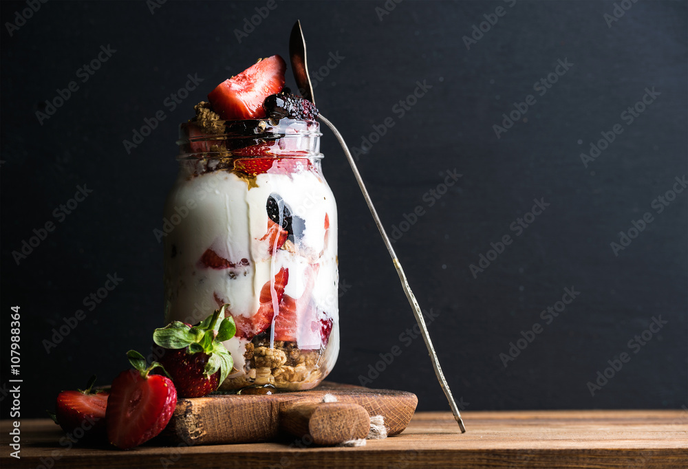 Yogurt oat granola with strawberries, mulberries, honey and mint leaves in tall glass jar on black b