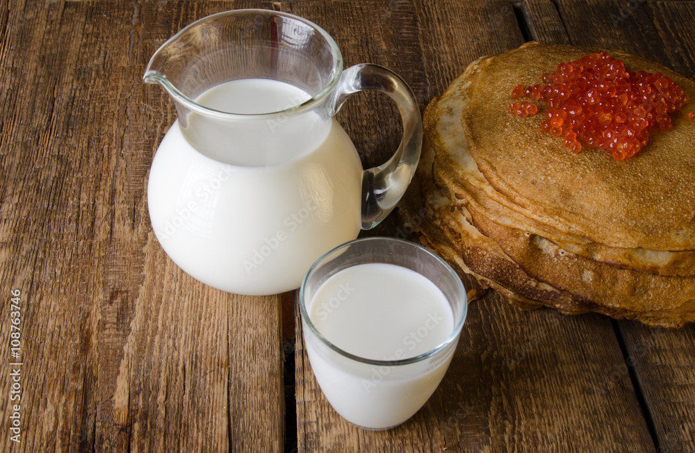 Glass jug and glass with milk on a wooden rustic background. Traditional Russian pancakes with red c