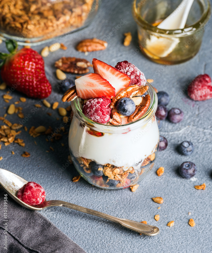 Yogurt oat granola with fresh berries, nuts, honey and mint leaves in glass jar on grey concrete tex