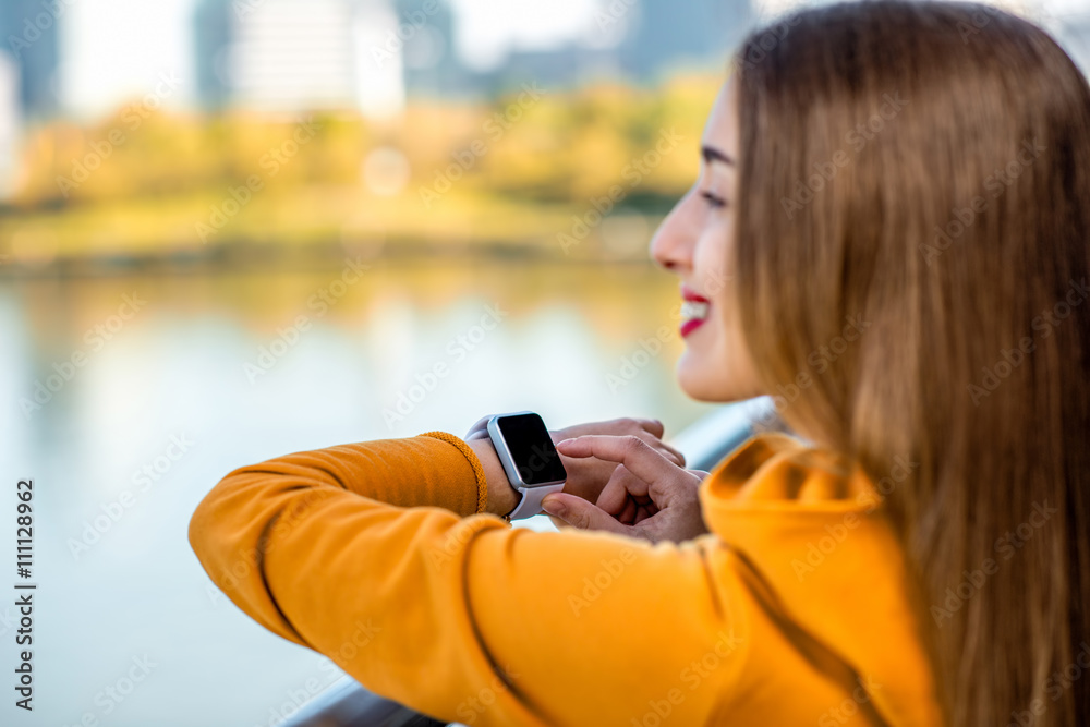 Young woman in yellow sweater using smart watch on the modern bridge with skyscrapers on the backgro