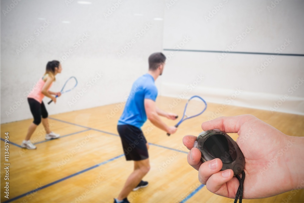 Composite image of coach is holding a stopwatch against couple enjoying a game of squash