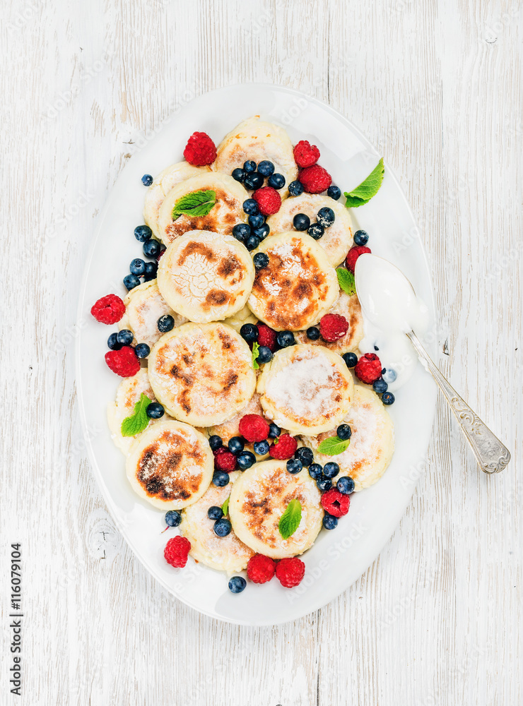 Syrniki or cottage cheese pancakes with fresh forest berries and sour cream sauce in serving dish ov