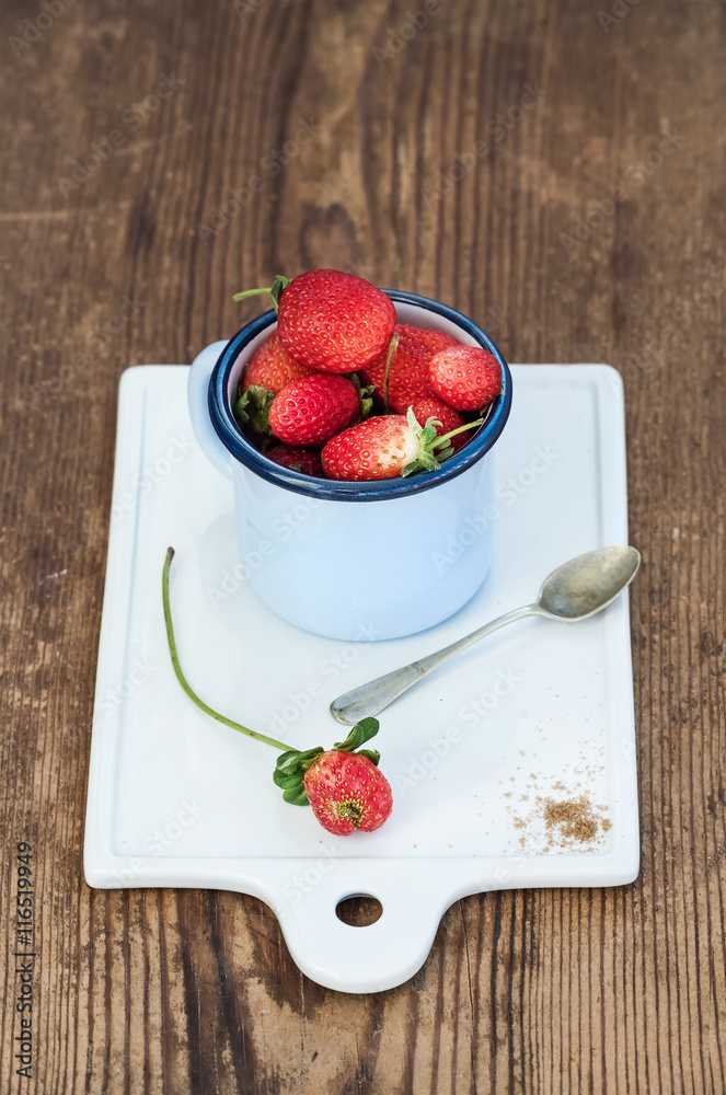 Fresh ripe red strawberries in blue enamel mug on white ceramic board over rustic wooden background,