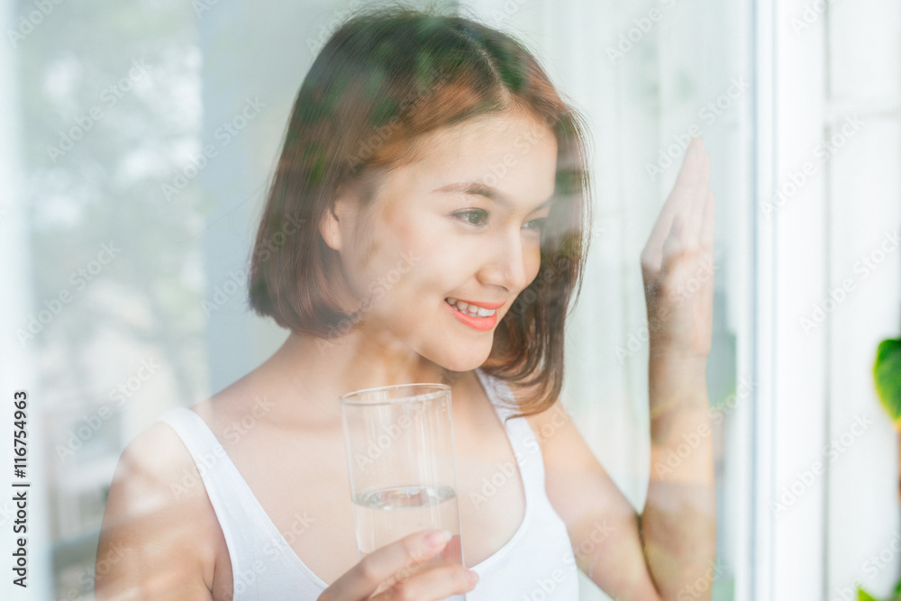 喝着水的年轻亚洲女人透过窗户看