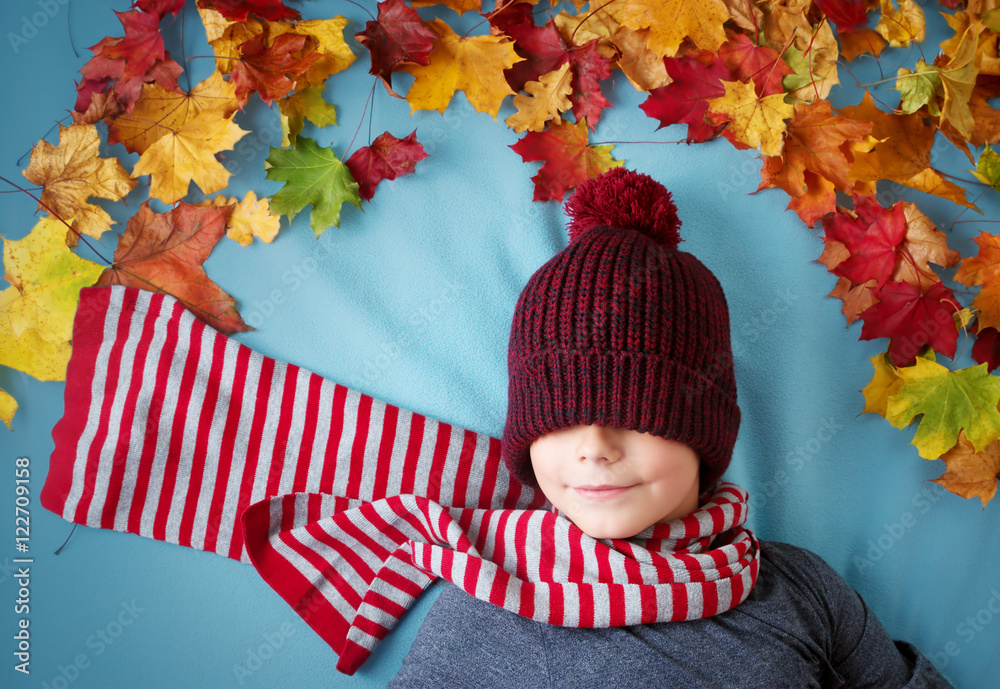 seven years old boy dreaming in autumn. Child in hat and scarf and maple leaves on blue background