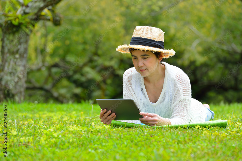 Happy middle aged woman lying on green grass using tablet computer in the park