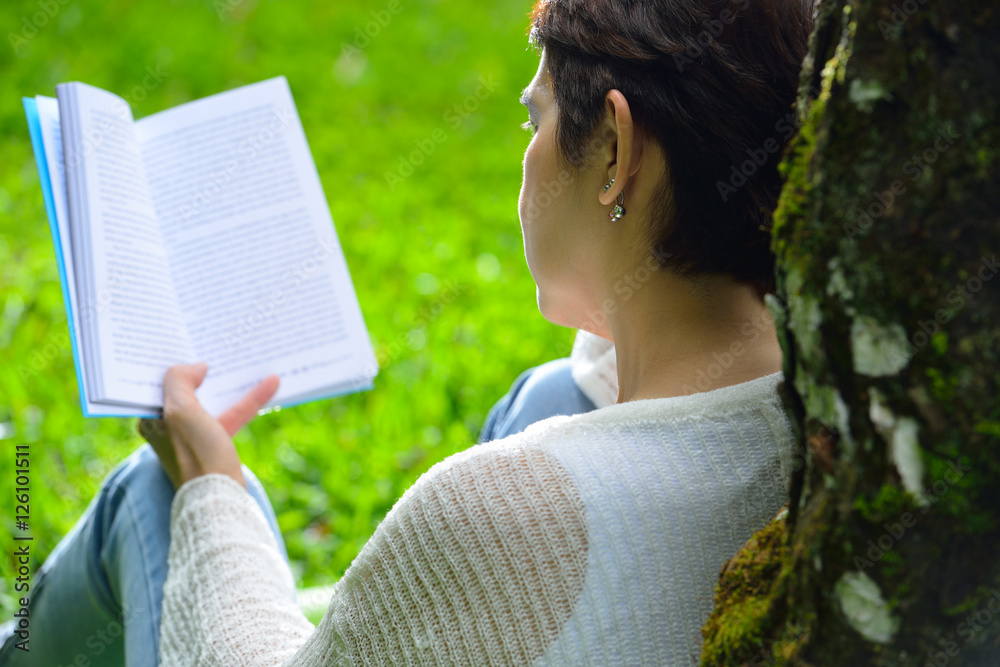 Middle aged woman sitting under a tree reading a book in the park