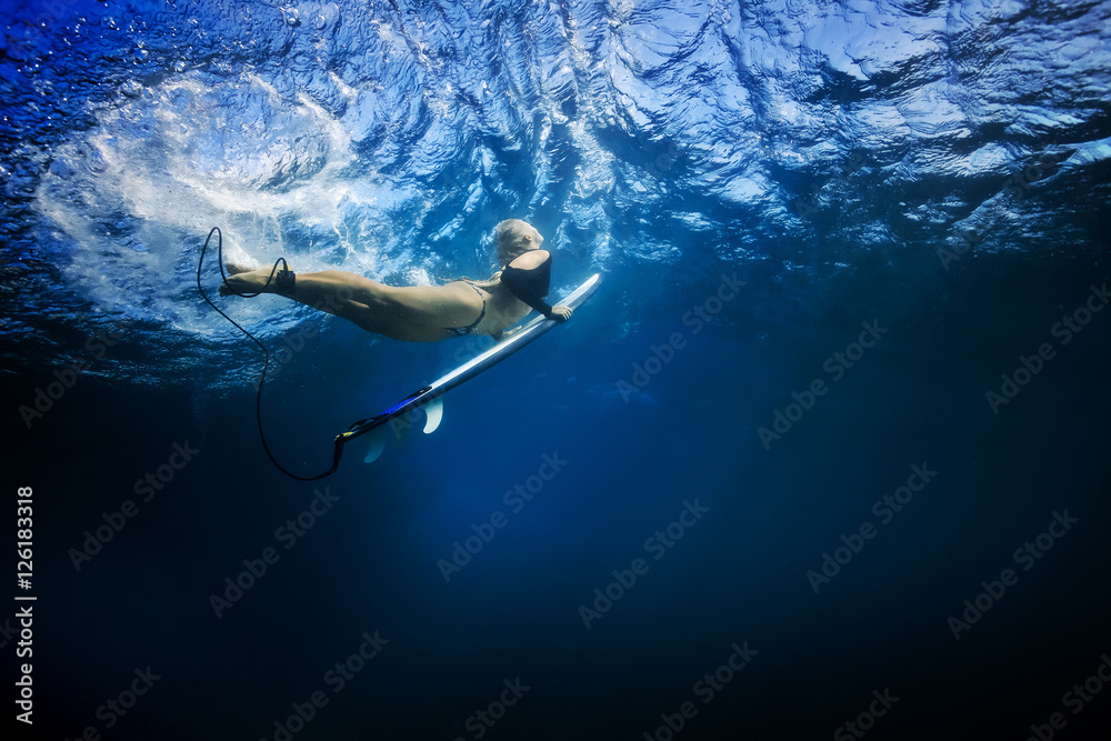 Slim blonde girl in bikini a surfer with surf board duck diving underwater under ocean reflected sur