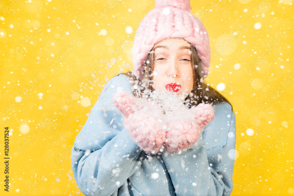 戴着针织帽子和手套的年轻女子站在黄色的墙上吹雪。Colorfu