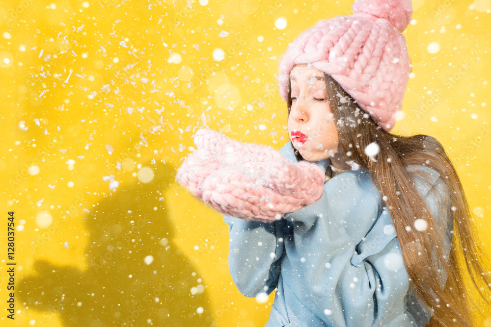 戴着针织帽子和手套的年轻女子站在黄色的墙上吹雪。Colorfu