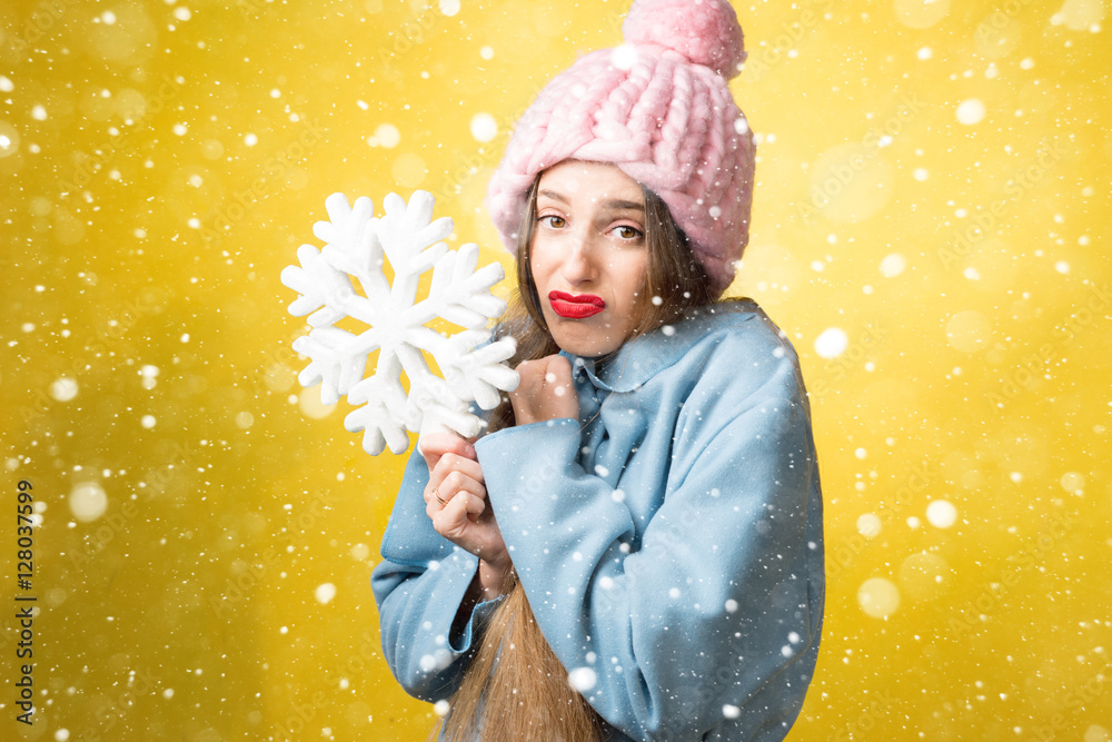 困惑而不快乐的女人穿着五颜六色的冬装，黄色背景上拿着一片雪花