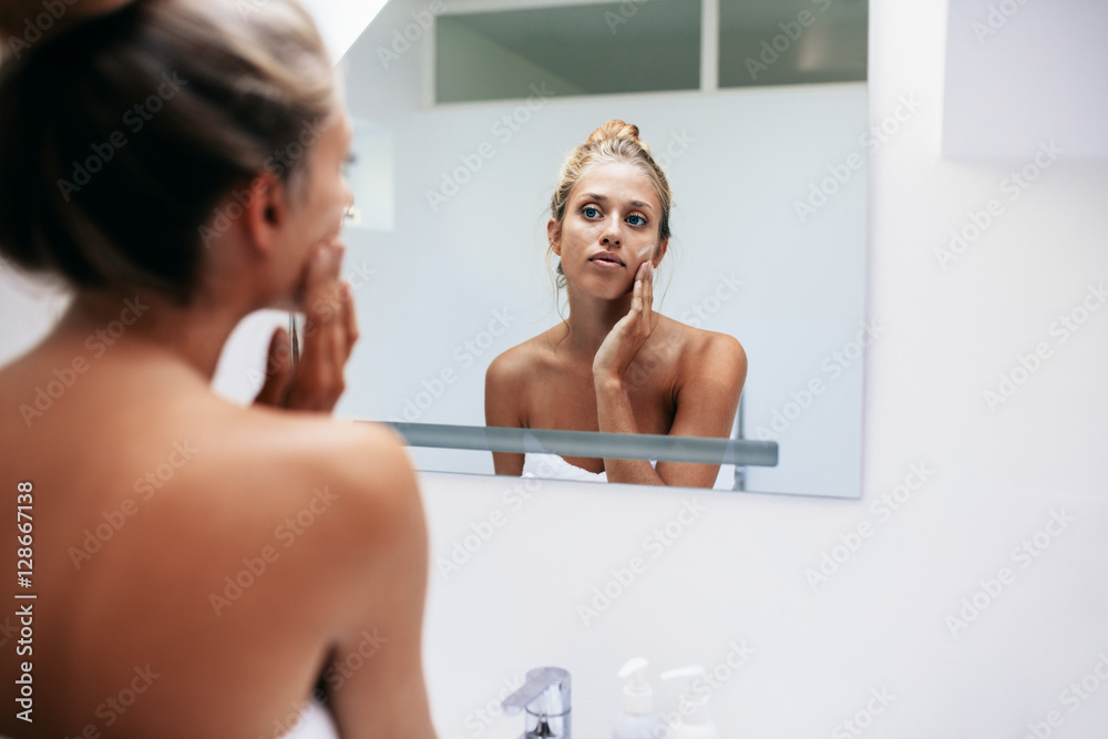 Beautiful woman applying moisturizer on face in bathroom