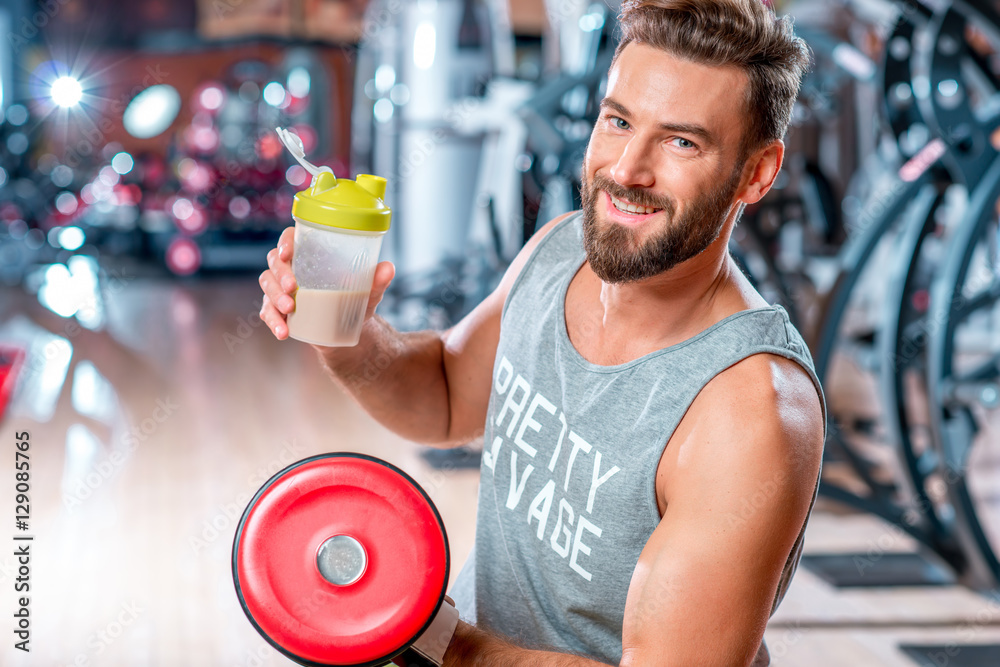 Muscular man drinking sports nutrition sitting with dumbbell in the gym