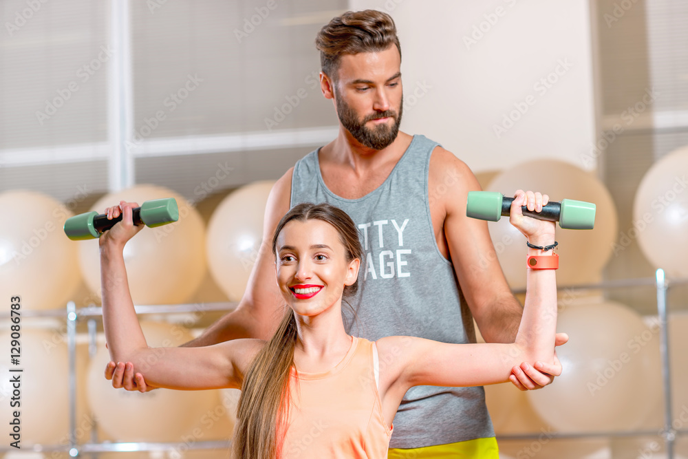 Young woman lifting dumbbells with personal trainer in the fitness room with fitballs on the backgro