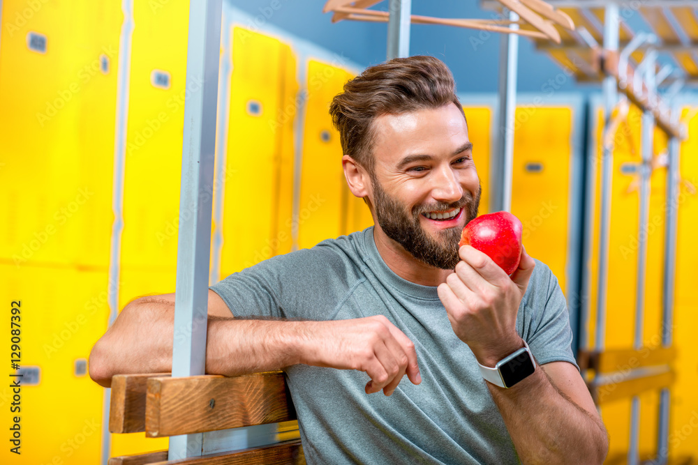 Sports man eating an apple sitting after the training in the locker room of the gym. Healthy natural