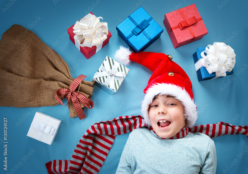 Happy child on blue blanket in red hat holding gift box. Cute boy in christmas cap with many present